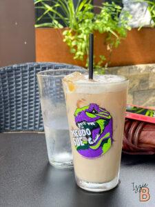 Ice-cold coffee drink in glass with Pseudo Sue logo, outdoor cafe scene, summer vibe.