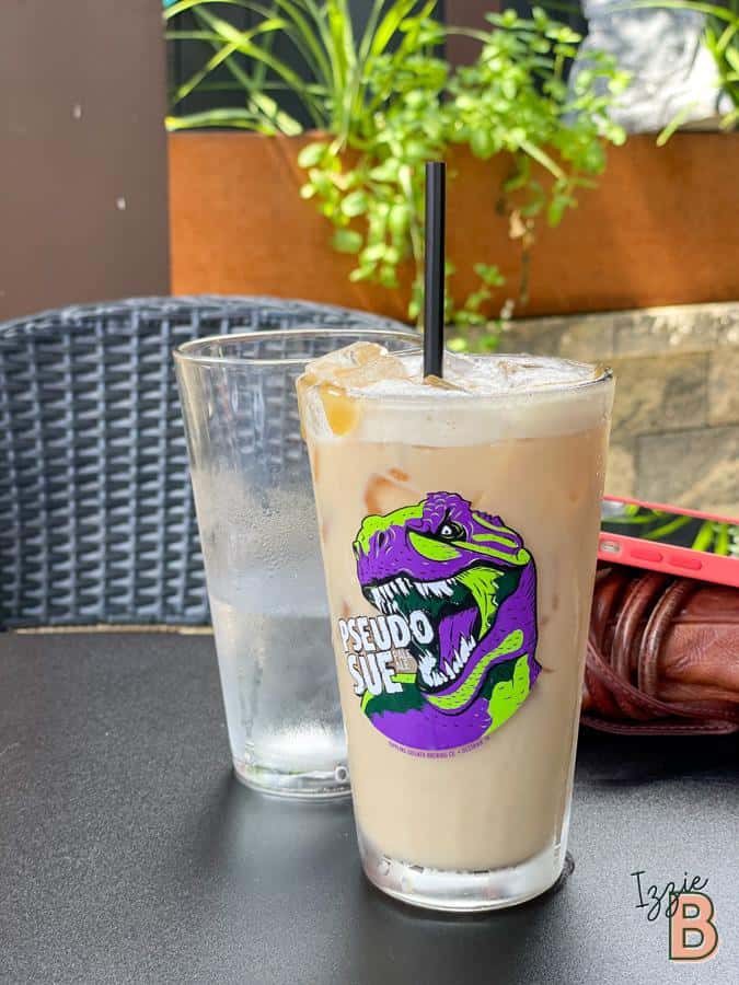 Ice-cold coffee drink in glass with Pseudo Sue logo, outdoor cafe scene, summer vibe.